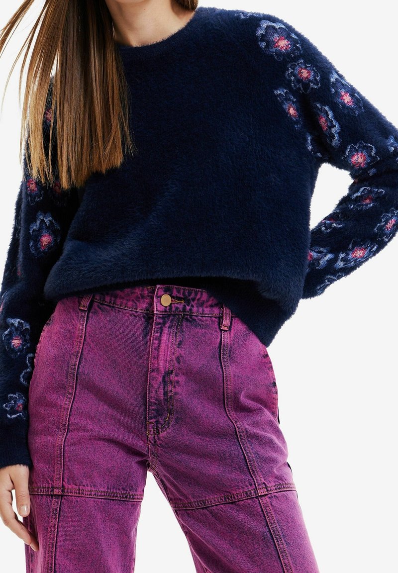 Femme portant un pull duveteux bleu marine avec des motifs floraux sur les manches et un jean violet vif taille haute, main reposant sur le côté.