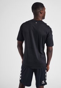 Man som vänder ryggen till i svarta sportkläder, inklusive en kortärmad tröja med en liten vit logotyp och matchande shorts med vita chevron-ränder.