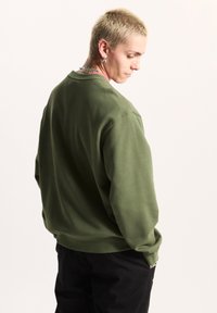 Felpa verde realizzata in misto cotone, caratterizzata da un classico collo a giro, vestibilità rilassata e polsini a costine. I pantaloni neri completano l'outfit.