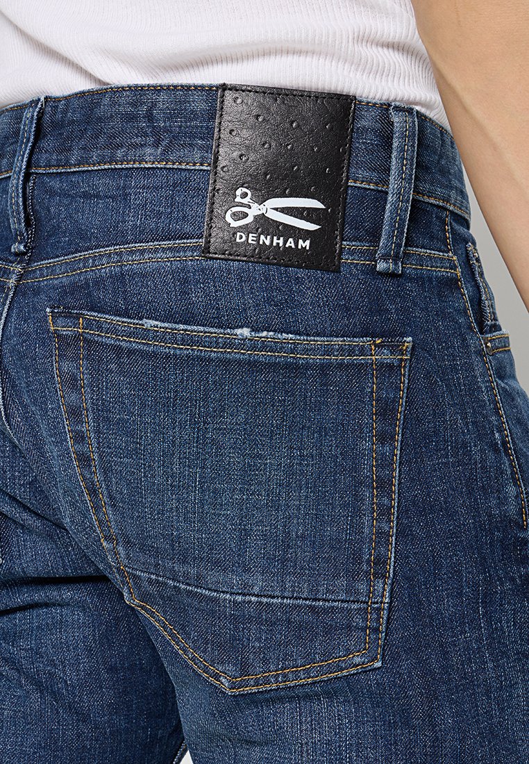 Donkere spijkerbroeken van denim met een gladde textuur. Beschikt over een zwarte leren patch met een reliëf van een schaarlogo, gele stiksels en details op de achterzak.