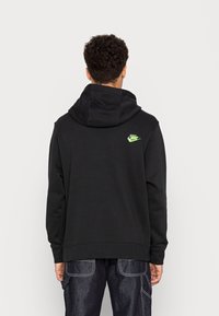 Svart hoodie i bomull med en grön Nike-logga på ryggen. Den har en avslappnad passform och en rymlig huva. Synliga sömdetaljer.