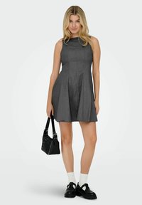 Giovane donna che indossa un vestito senza maniche grigio scuro, calze bianche, scarpe nere chunky, mentre tiene una borsa nera, in piedi contro uno sfondo semplice.