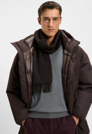 Brun pufferjakke med skinnende finish, grå sweater og mørk tørklæde med frynsedetaljer. Tørklædet har et lille logo-applikation.