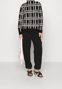 Un pull à motifs noir et gris avec un col haut, associé à un pantalon noir ample et des sandales noires à lanières avec des accents jaunes. Sac à main rose clair.