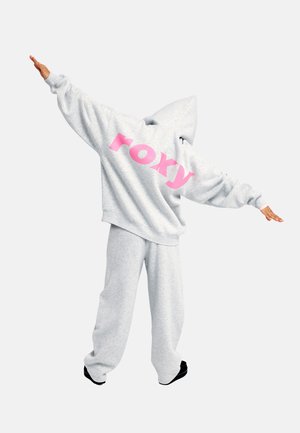 Person trägt grauen Hoodie und Jogginghose mit weit ausgebreiteten Armen, auf dem Hoodie steht großer pinker "roxy"-Text auf dem Rücken.