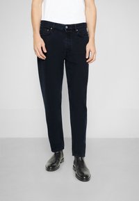 Man i smala mörkblå jeans, vit t-shirt och svarta läderanstickade stövlar, stående mot en plain ljus bakgrund.