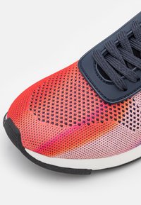 Chaussure de sport en maille avec un dégradé de rouge et rose, lacets bleu marine foncé et semelle en caoutchouc texturée.