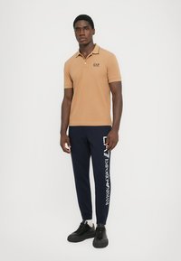 Brown polo shirt with black collar and EA7 logo, paired with navy joggers featuring white Emporio Armani text, and black sneakers.