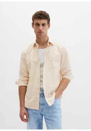 Νέος άνδρας που φοράει μια μπεζ ριγέ πουκάμισο με κουμπιά πάνω από λευκή φανέλα και ανοιχτό μπλε τζιν, στέκεται με το ένα χέρι στην τσέπη.
