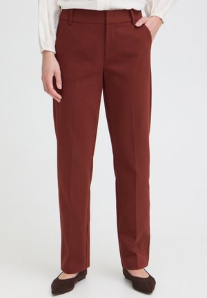 Personne portant un pantalon droit couleur rouille, une blouse blanche, des ballerines brunes ; une main dans la poche, debout devant un fond uni.
