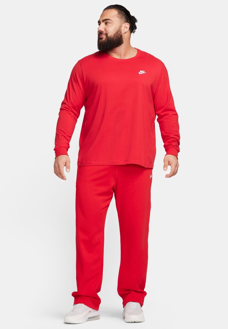 Czerwona koszula z długim rękawem i pasujące spodnie wykonane z mieszanki bawełny. Posiadają białe logo Nike na klatce piersiowej i nodze. Obuwie jest w jasnym kolorze.