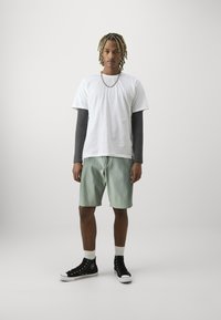 Camiseta de algodón blanca superpuesta sobre una camisa de manga larga gris, combinada con pantalones cortos verde claro y zapatillas altas negras con suelas blancas.