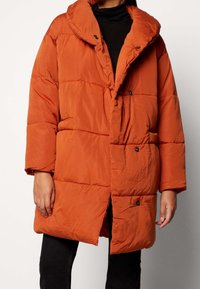 Vadderad orange kappa med huva, med knappstängning fram, sidofickor och en avslappnad passform. Slät textur och quiltad design.