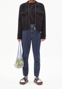 Schwarze, kürzere Jacke mit zwei vorderen Taschen, kombiniert mit hochgeschnittenen blauen Jeans. Das Modell hält eine Netztragetasche mit Gemüse und trägt schwarz-weiße Sandalen.