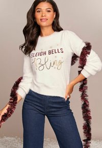 Maglione bianco e soffice con la scritta "SLEIGH BELLS BLING" in glitter rosso borgogna e oro. Rifinito con lustrini borgogna lungo le maniche. Indossati jeans scuri.