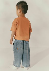 Īsām piedurknēm apelsīnu krāsas kokvilnas T-krekls, kas saderē ar platām zilu džinsu biksēm ar divām aizmugurējām kabatām un smaidošas sejas detaļu. Baltas kurpes.