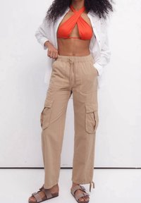 Pantalons cargo beiges avec une taille élastique et des poches latérales, assortis à un haut de bikini halter orange vif et une chemise blanche à boutons.