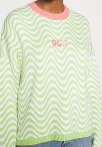 Jasnozielony i biały sweter z falistym wzorem, różowym ściągaczem przy szyi oraz pomarańczowym, haftowanym logo "RAGGED JEANS" z przodu.