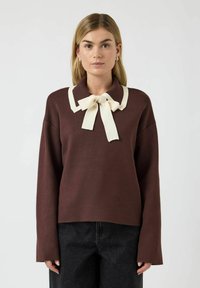 Brun genser med lange ermer, kremfarget krage og sløyfedetalj. Har en avslappet passform og myk tekstur. Bært med mørke jeans.