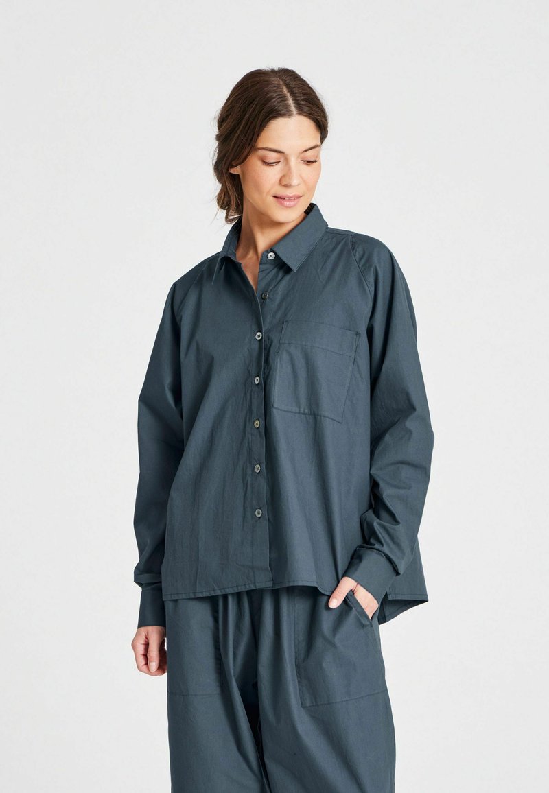 Camicia button-up verde scuro con colletto, maniche lunghe e tasca sul petto, indossata con pantaloni larghi abbinati.