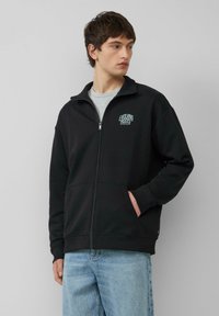Zwarte zip-up sweatshirt met hoge kraag, voorzien van een klein wit logo op de borst. Zachte textuur, twee voorzakken en geribde manchetten.