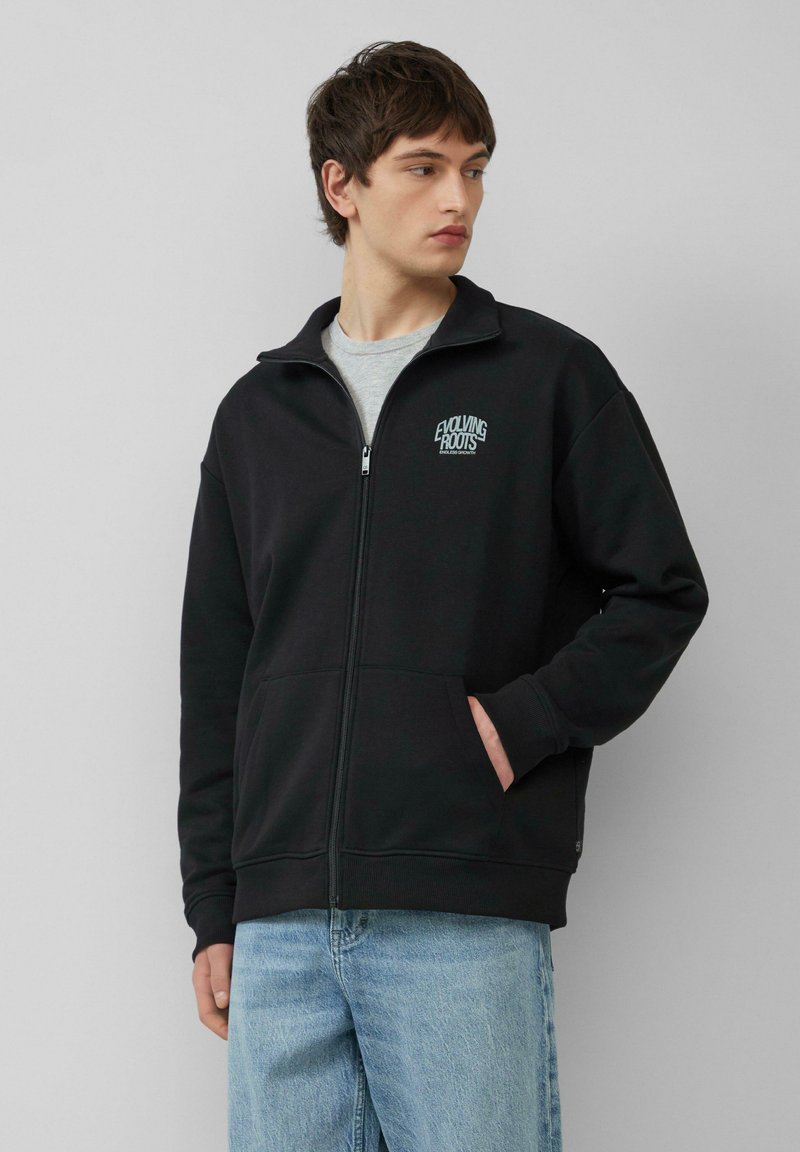 Zwarte zip-up sweatshirt met hoge kraag, voorzien van een klein wit logo op de borst. Zachte textuur, twee voorzakken en geribde manchetten.