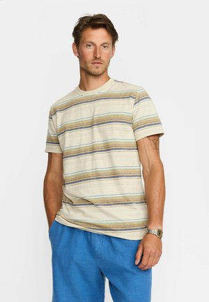 Mand iført beige t-shirt med striber og blå shorts, stående med den ene hånd i lommen mod en ensfarvet baggrund.