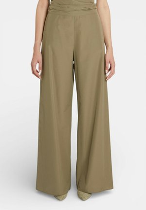 Pantaloni verde oliva a gamba larga indossati da una persona in piedi, abbinati a scarpe beige con punta appuntita, su uno sfondo bianco semplice.