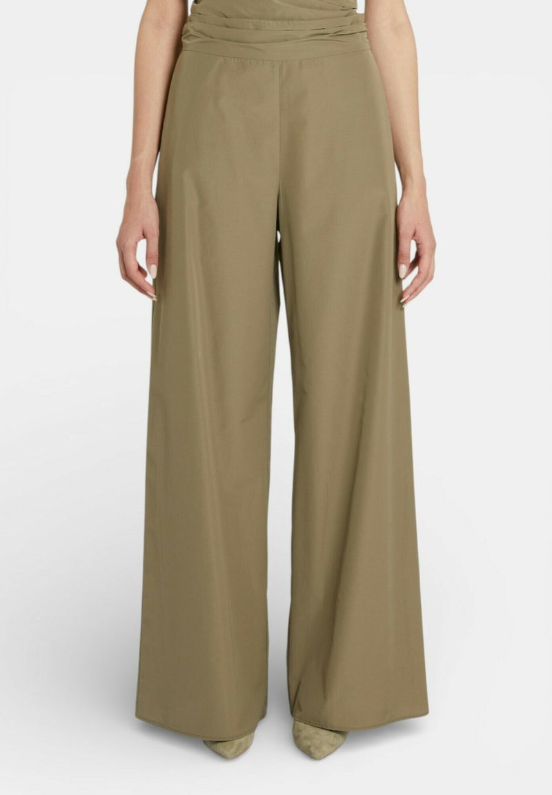 Pantaloni verde oliva a gamba larga indossati da una persona in piedi, abbinati a scarpe beige con punta appuntita, su uno sfondo bianco semplice.