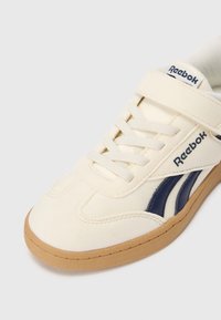 Off-white Reebok tenisky s modrými logovými pruhmi, béžovou podrážkou, šnúrami a uzáverom na suchý zips na bielom pozadí.