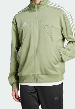 Persoon die een olijfgroene Adidas jas met ritssluiting draagt met witte strepen op de mouwen en zwarte Adidas broek, hand in de jaszak.