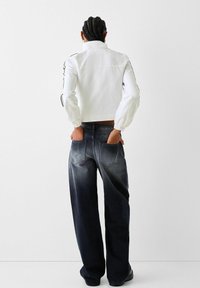 Krótkia biała kurtka z elastycznymi mankietami, zestawiona z jeansami w ciemnym kolorze o wysokim stanie i szerokich nogawkach, z efektem wyblakłego gradientu i tylnymi kieszeniami.