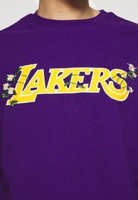 Fialové bavlněné tričko s velkým žlutým logem "LAKERS", zdobené květinovou výšivkou a zelenými výhonky kolem písmen.