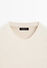 Maglione a coste off-white con colletto rotondo e un'etichetta nera cucita all'interno del collo.
