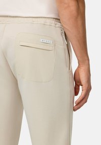 Beige trenirke izdelane iz mehkega materiala, z elastičnim pasom in zadnjim žepom z oznako. Gladka tekstura, ravna kroja.