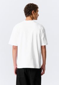 Homme aux cheveux courts et bouclés, portant un t-shirt blanc uni oversize et un pantalon noir, vu de dos sur un fond gris clair.