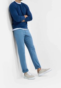 Modrý svetr, bílá košile, světle modré kalhoty a béžové tenisky s bílými podrážkami. Oblečení má uvolněný střih a ohrnuté rukávy.