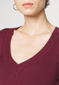 Vinröd ribbad tröja med djup V-halsringning, som har texturerad tyg och en enkel design som betonar en figursydd silhuett.