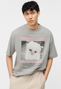 Young man wearing a grey t-shirt with a black-and-white missing cat poster and pink text offering a $1000 reward.