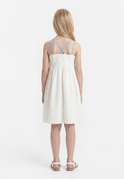 Dievča s dlhými blond vlasmi, oblečené v bielych bezrukávových šatách s prekríženými modrými ramienkami na chrbte a bielych sandáloch, stojí obrátené chrbtom.