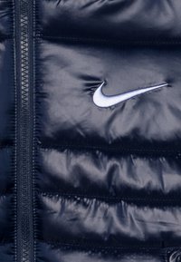 Námořnicky modrá prošívaná bunda s lesklým povrchem, vybavená celoplozným zipem a bílým vyšitým logem Nike na hrudi.