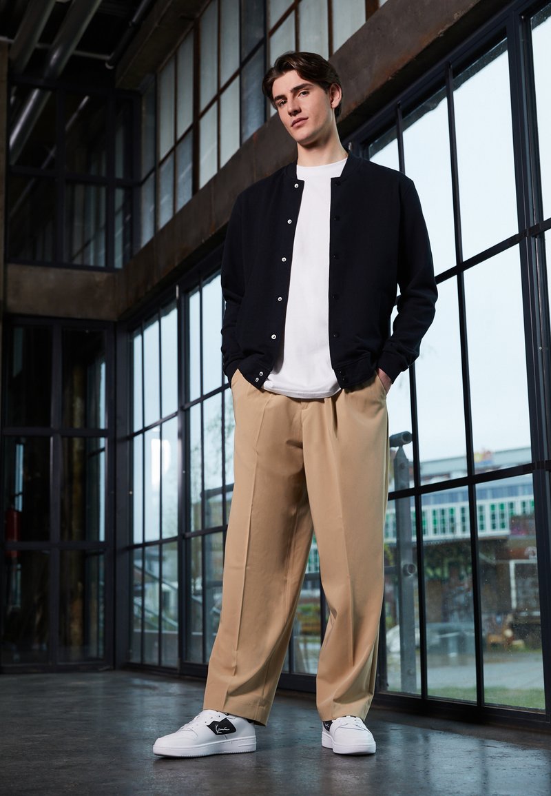 Svart knappet jakke over en hvit skjorte, paret med beige løse bukser og hvite joggesko med svarte detaljer, satt mot store glassvinduer.