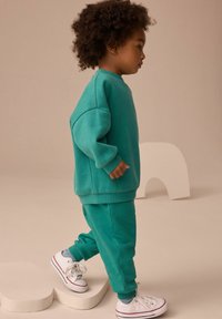 Turkos sweatshirt och matchande joggers, tillverkade av mjukt material, med ribbade ärmslut och vita sneakers med röda detaljer, stående på en skulptural bas.