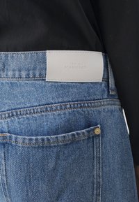 Světle modré džíny z denimu s hladkým povrchem, s bílou koženou visačkou s nápisem "7 FOR ALL MANKIND" na pase a kapsou se zlatým kováním.