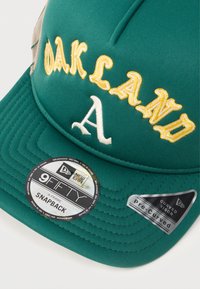 Zelena snapback kapa s izvezenim natpisom "OAKLAND" i "A." Ima zakrivenu vizir i teksturu tkanine, označena s New Era i 9FIFTY oznakama.
