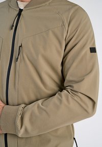 Giacca bomber beige con zip frontale, due tasche sul petto e polsini elastici. Realizzata in tessuto liscio con una finitura leggermente texture.