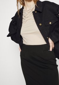 Chaqueta de mezclilla negra con botones dorados, top beige con cuello drapeado y falda lápiz verde oscuro con un bolsillo lateral. Texturas suaves, diseño casual.