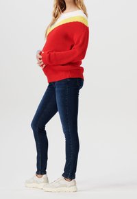 Raska kvinna i röd, gul och vit tröja, mörkblå jeans och vita sneakers, som håller om sin mage framför en enfärgad bakgrund.