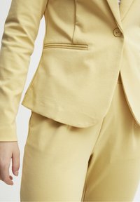 Blazer jaune clair à un seul bouton, col cranté et poche latérale ; associé à un pantalon assorti taillé sur mesure, texture de tissu lisse.