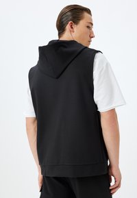 Hoodie nera senza maniche con cappuccio, realizzata in tessuto morbido, abbinata a una maglietta bianca a maniche corte. Presenta una vestibilità comoda e orli a costine.